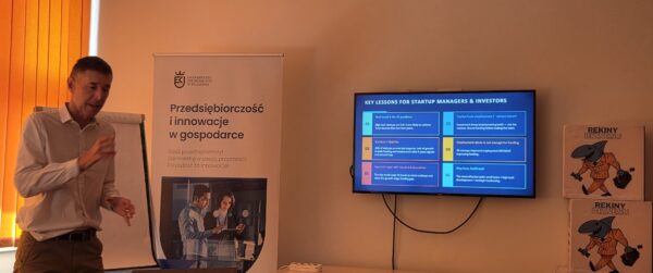 Prowadzący w sali, obok plakat kierunku Przedsiębiorczość i ekran z przezentacją
