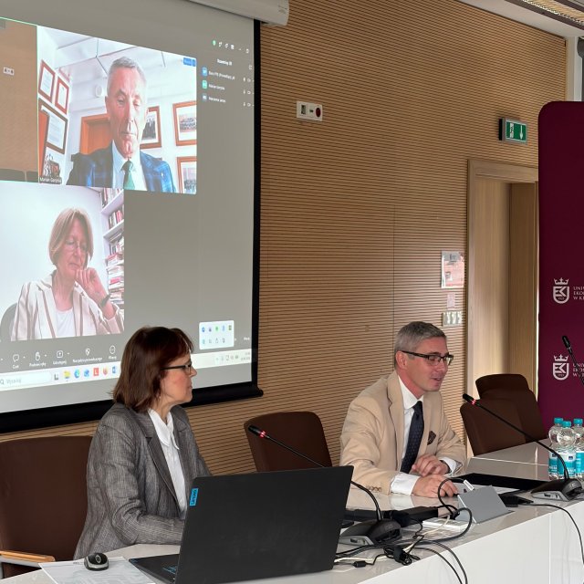 Widok na st&oacute;ł prezydialny przy kt&oacute;rym siedzą prof. M. Urbaniec i prof. M. Folwarski, w tle obrazy uczestnik&oacute;w on-line