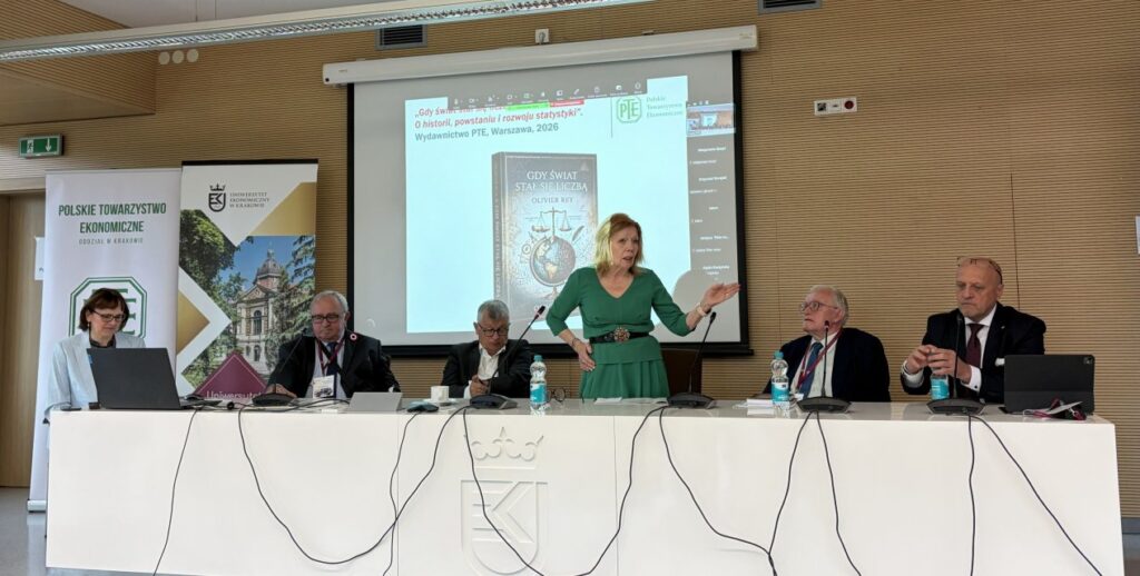 Zdjęcie panelist&oacute;w - 3 osoby za stołem siedzą, pośrodku prelegentaka stoi przed swoją prezentacją